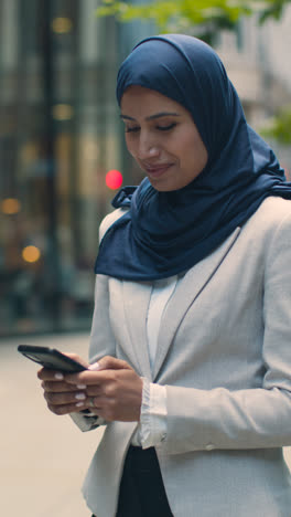 Vertikales-Video-Einer-Muslimischen-Geschäftsfrau,-Die-Nachrichten-Auf-Dem-Mobiltelefon-überprüft,-Während-Sie-Vor-Dem-Büro-In-Stadt-1-Steht